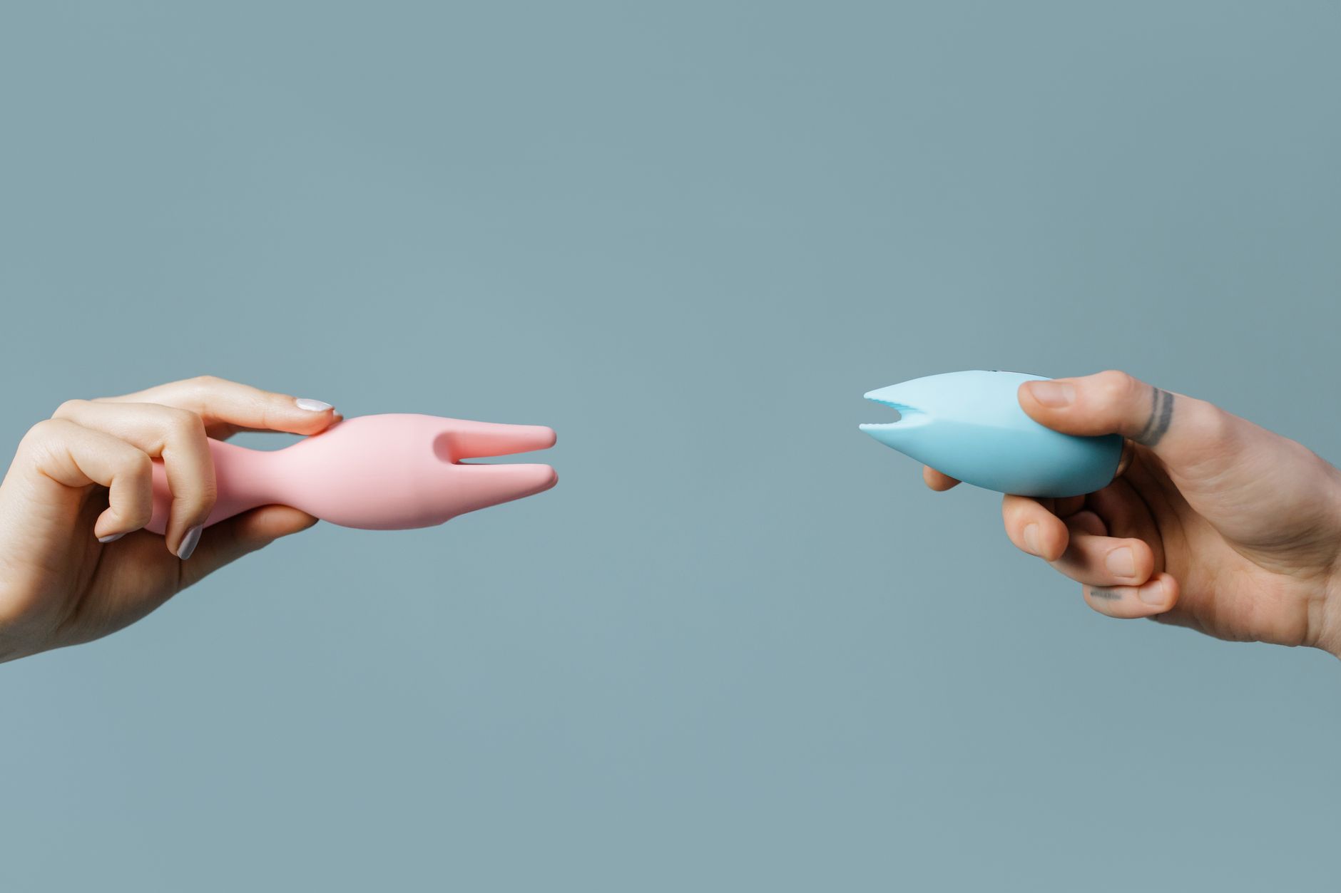 Two hands holding pink and blue silicone vibrators against a pastel background, representing shared intimacy and reconnection.