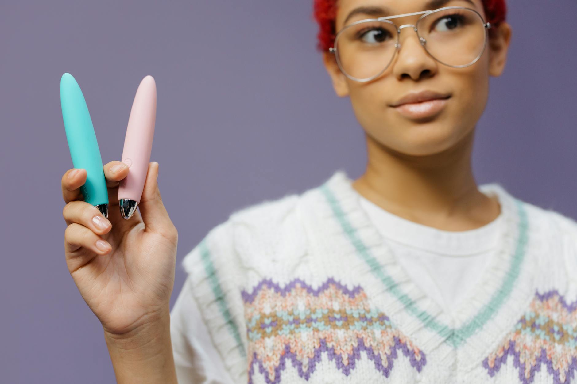 Une femme avec des lunettes tient deux vibromasseurs colorés contre un fond violet.
