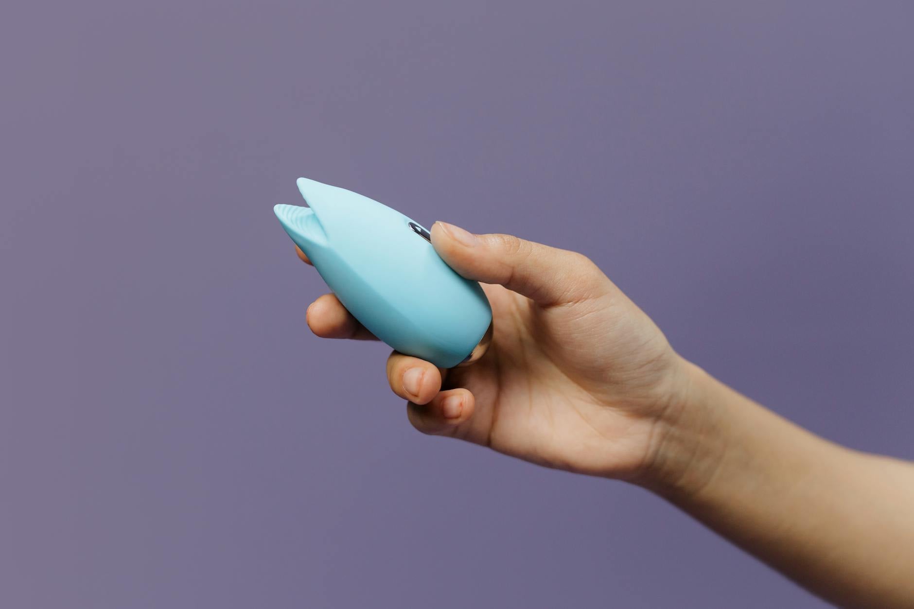 A blue silicone clitoral vibrator held in hand against a purple background, symbolizing self-pleasure and intimacy after surgery