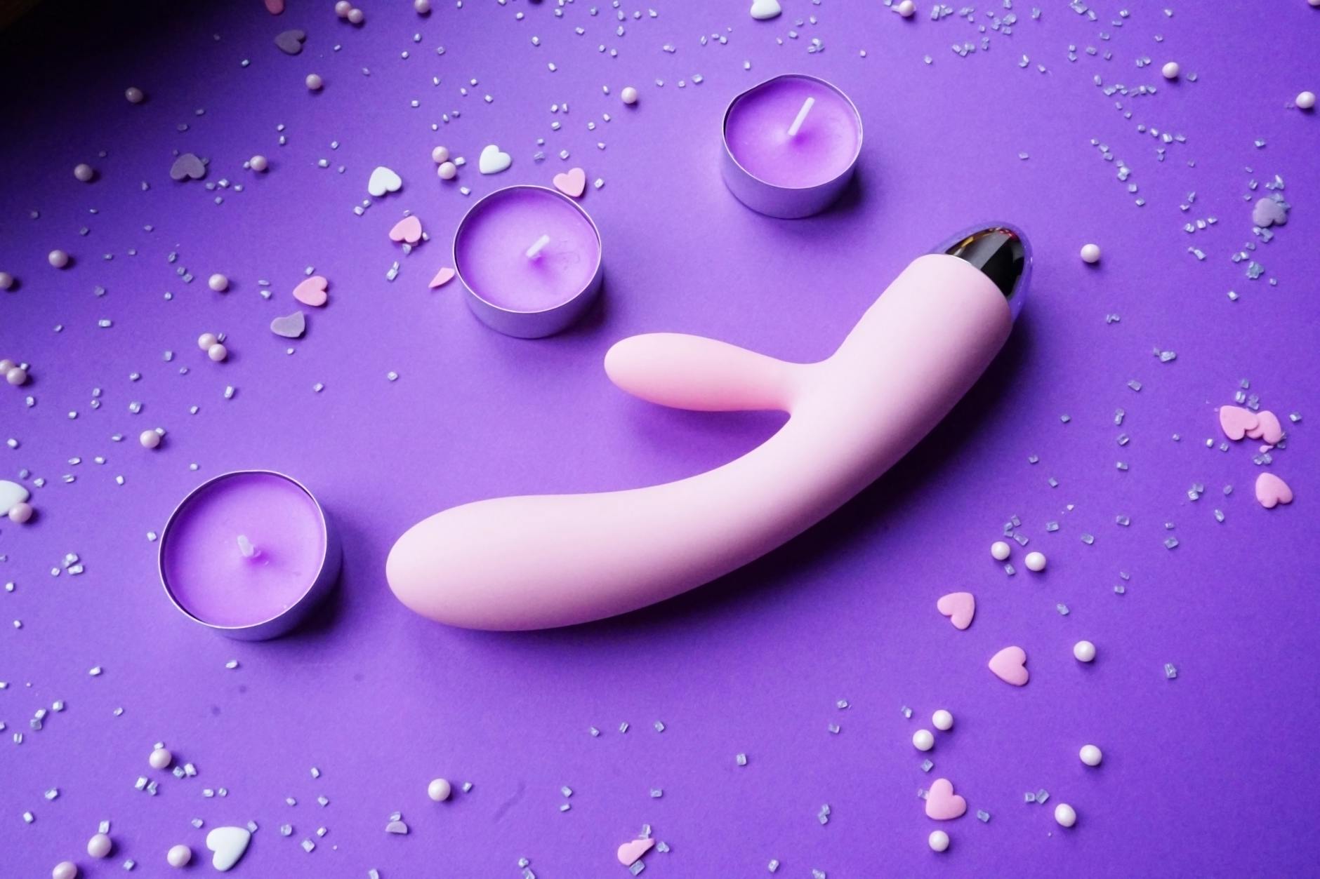 A vibrator on a purple background with heart confetti and candles for a romantic vibe.